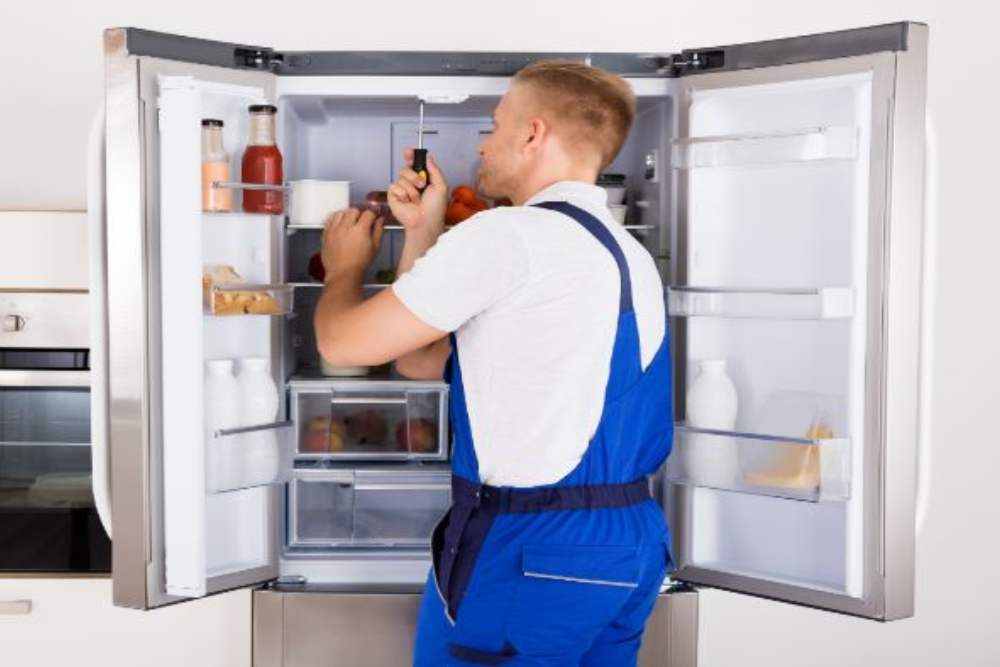 A refrigerator technician in uniform checking and repairing a fridge in a kitchen.