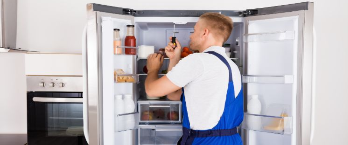 A refrigerator technician in uniform checking and repairing a fridge in a kitchen.